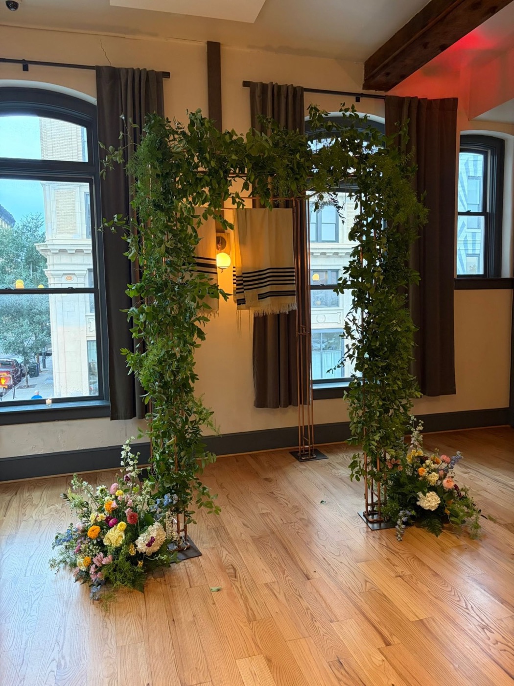 Copper chuppah wrapped in greenery with a tallit canopy and wildflowers at the base, in a loft with arched windows